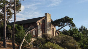 Merrill Hall at Asilomar, Julia Morgan's final building on the grounds, its native stone chimney and timber gable rising among Monterey pines in late afternoon light